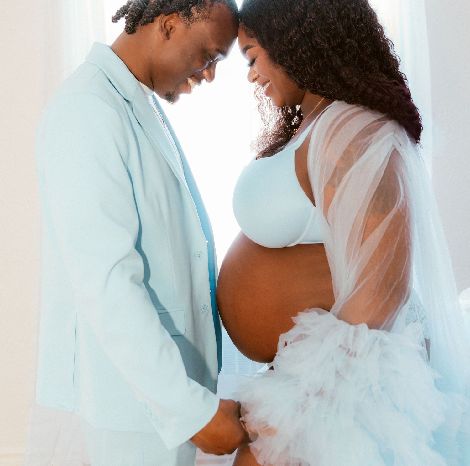 Romantic maternity photo shows a couple in light blue and white sharing an intimate moment in a bright setting.
