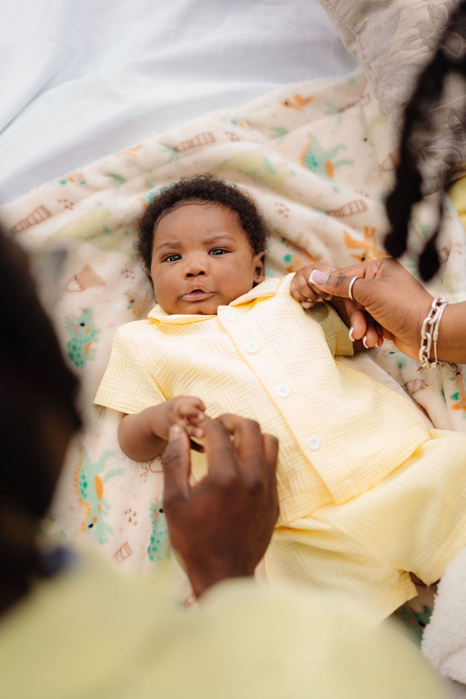 A baby wearing a yellow onesie is being gently dressed by adult hands on a soft white blanket.