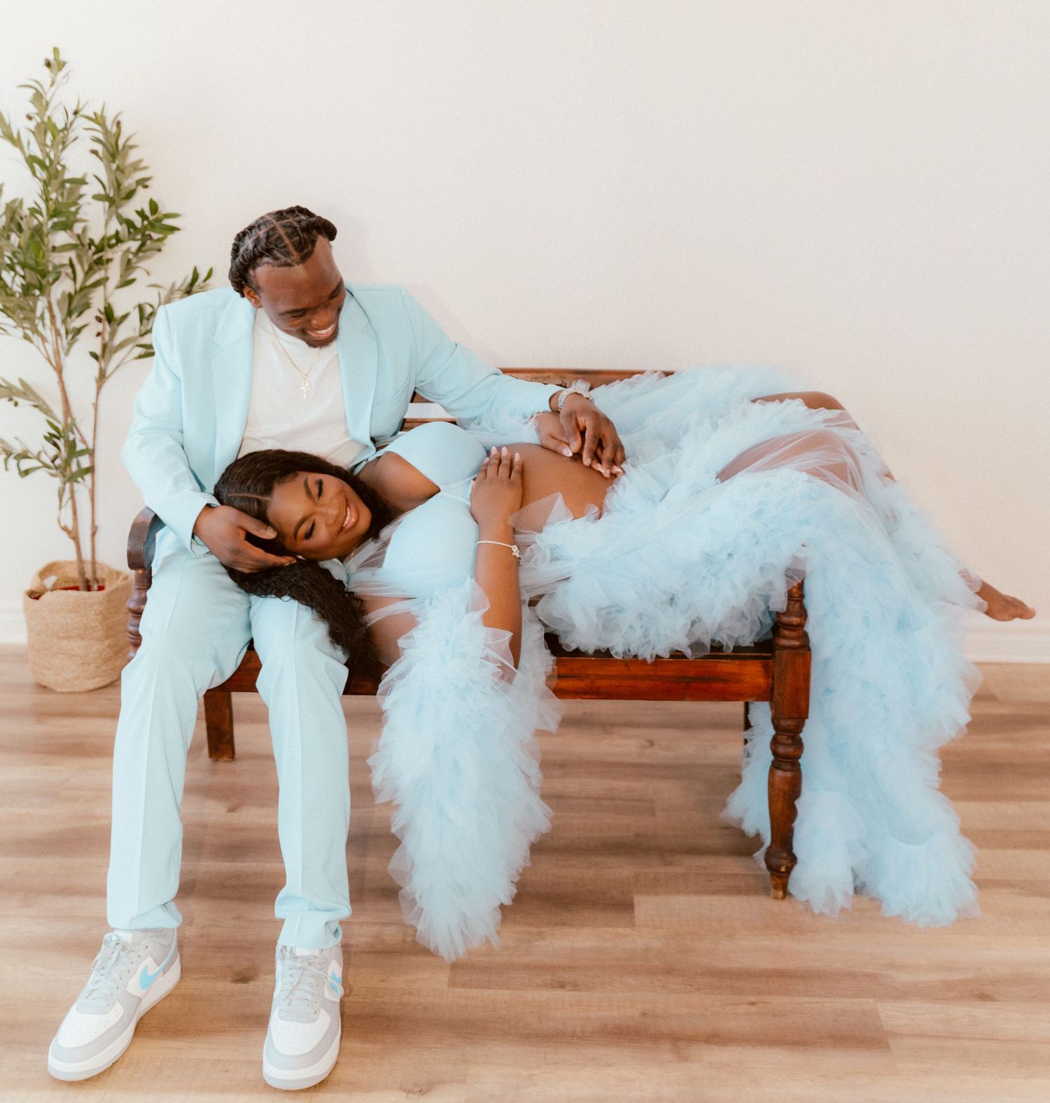 Two people in matching light blue outfits pose together on a wooden bench with tulle details and a potted plant nearby.