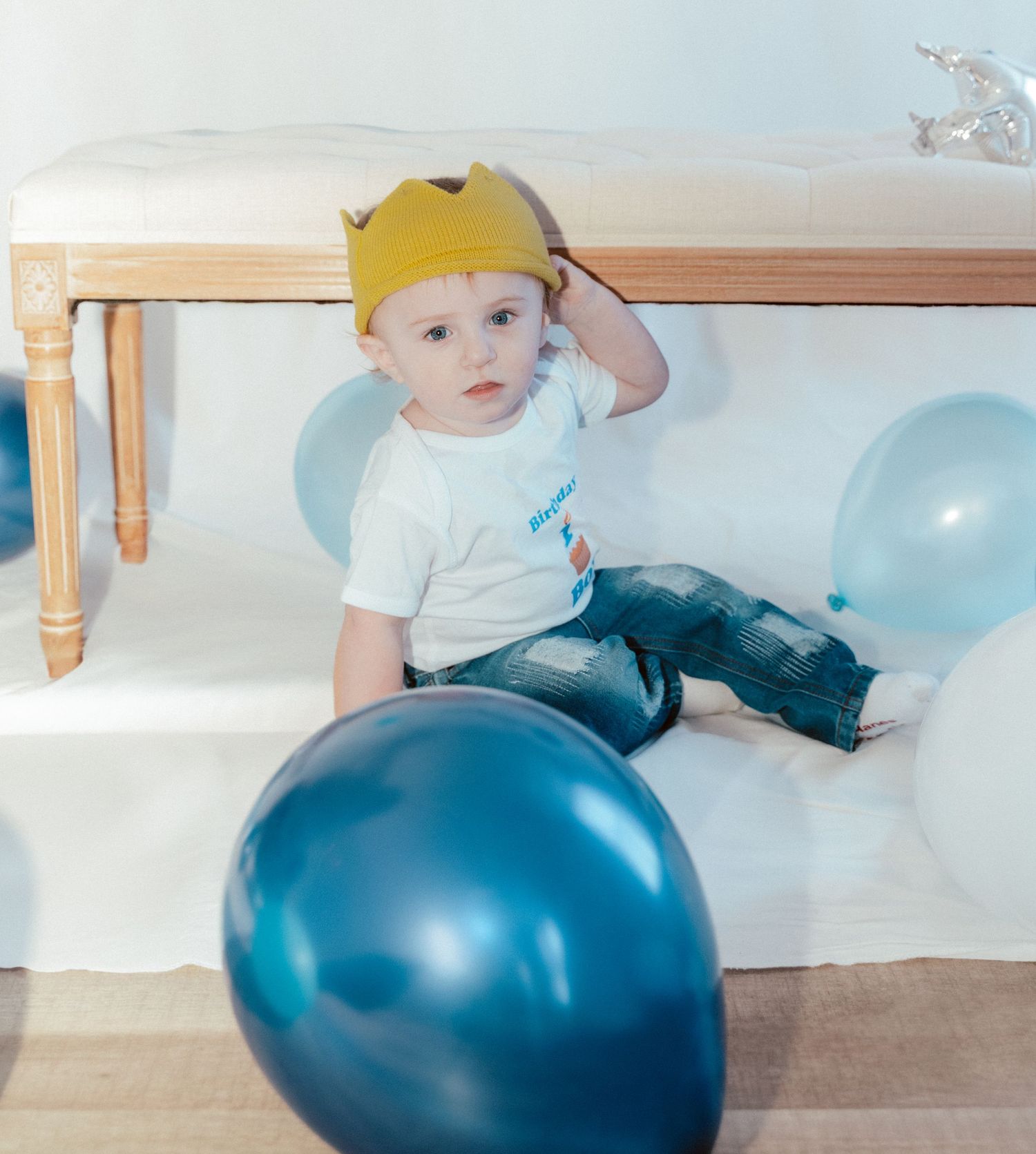 A small figure in a yellow hat sits on a floor with blue balloons during a celebration.