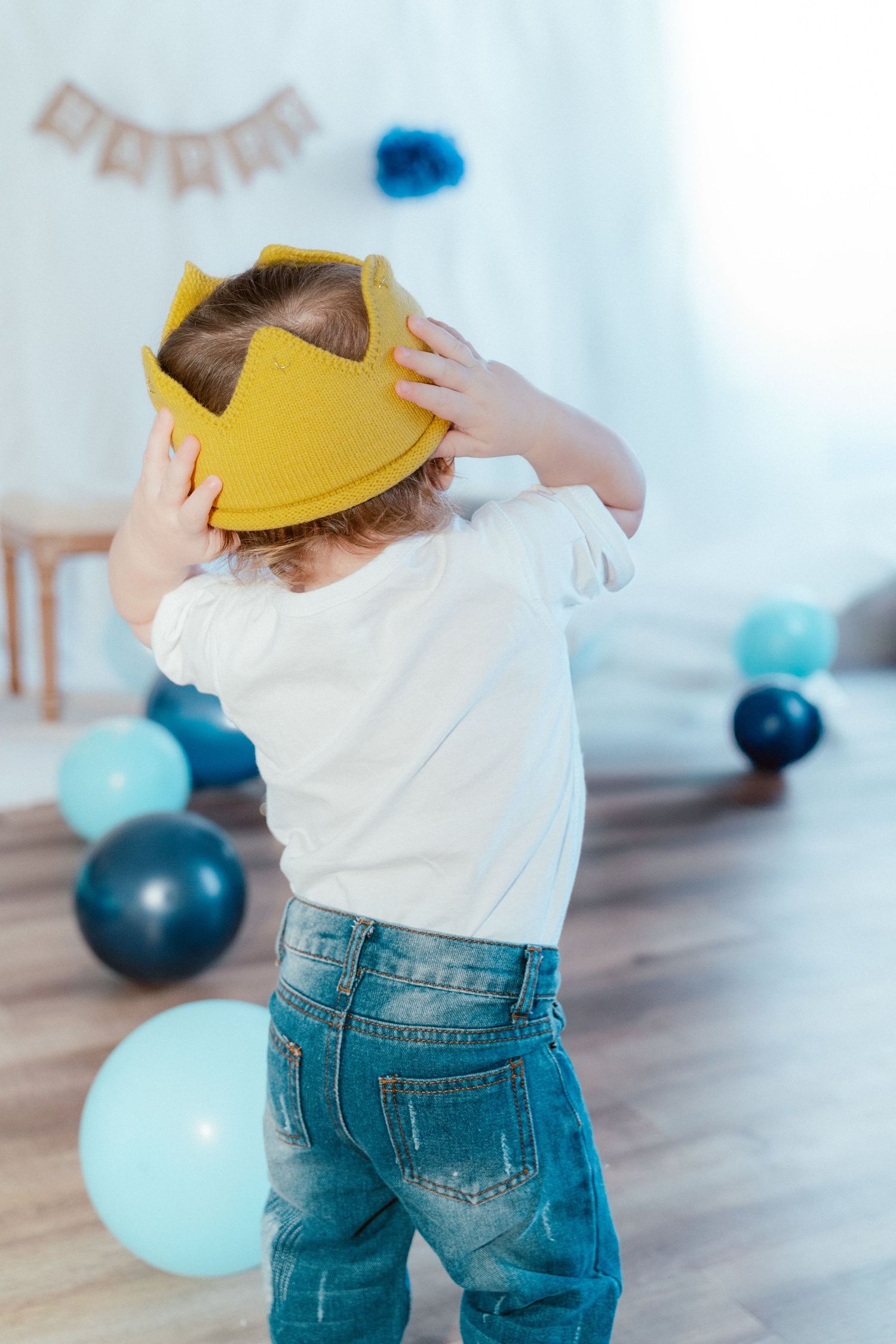 Small child wearing a yellow paper crown and jeans dances among blue balloons at a party.