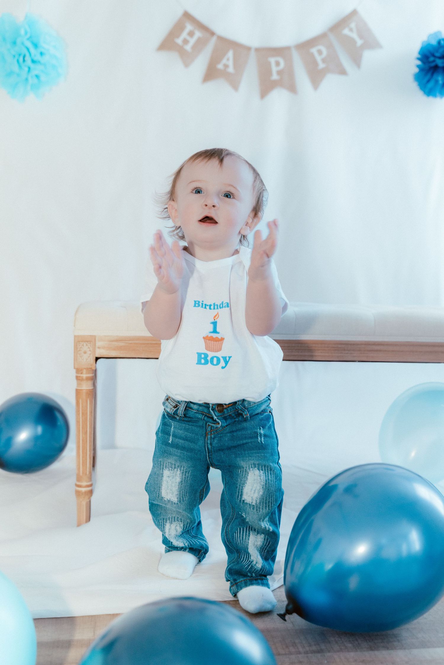 Baby wearing a 'Boy' shirt and jeans sits next to blue balloons and birthday decorations at a first birthday party.