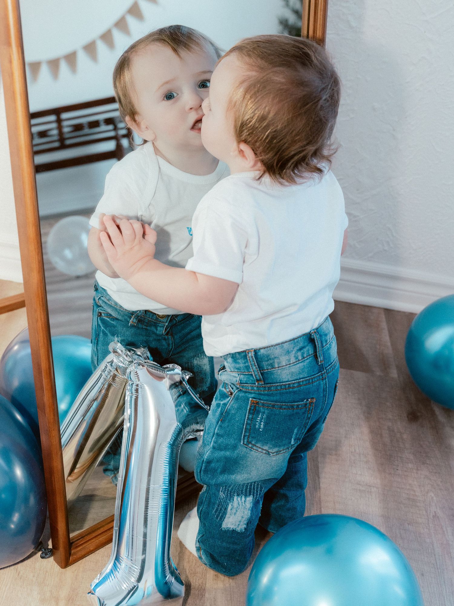 A toddler in white shirt and jeans looks at their reflection in a full-length mirror with blue balloons and party decorations.