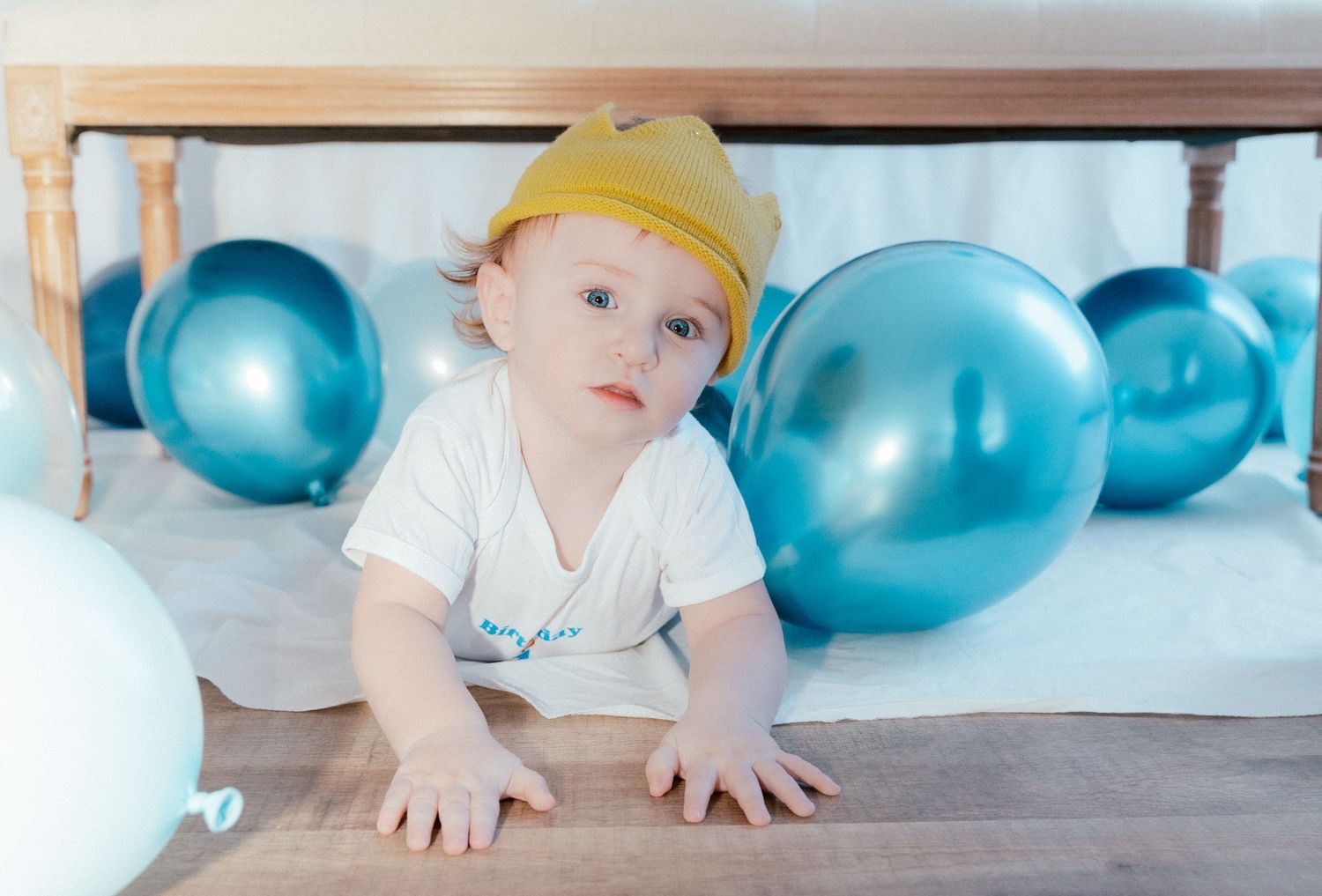 Small child in yellow hat crawls under table with blue balloons on wooden floor.