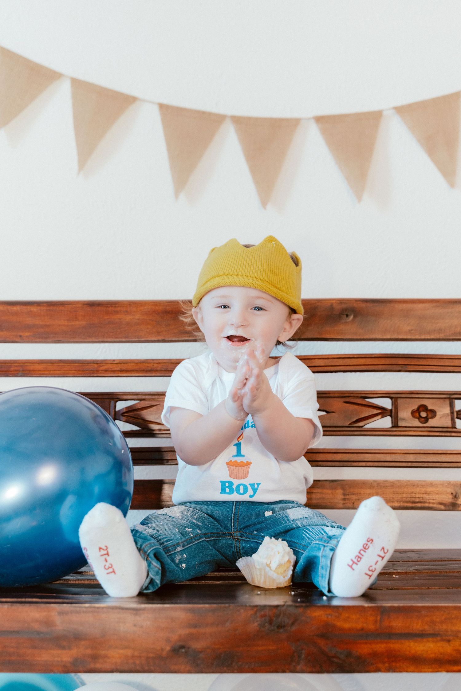 Baby in a yellow crown and "Boy" t-shirt sits on wooden bench with blue balloons for birthday celebration photo session.