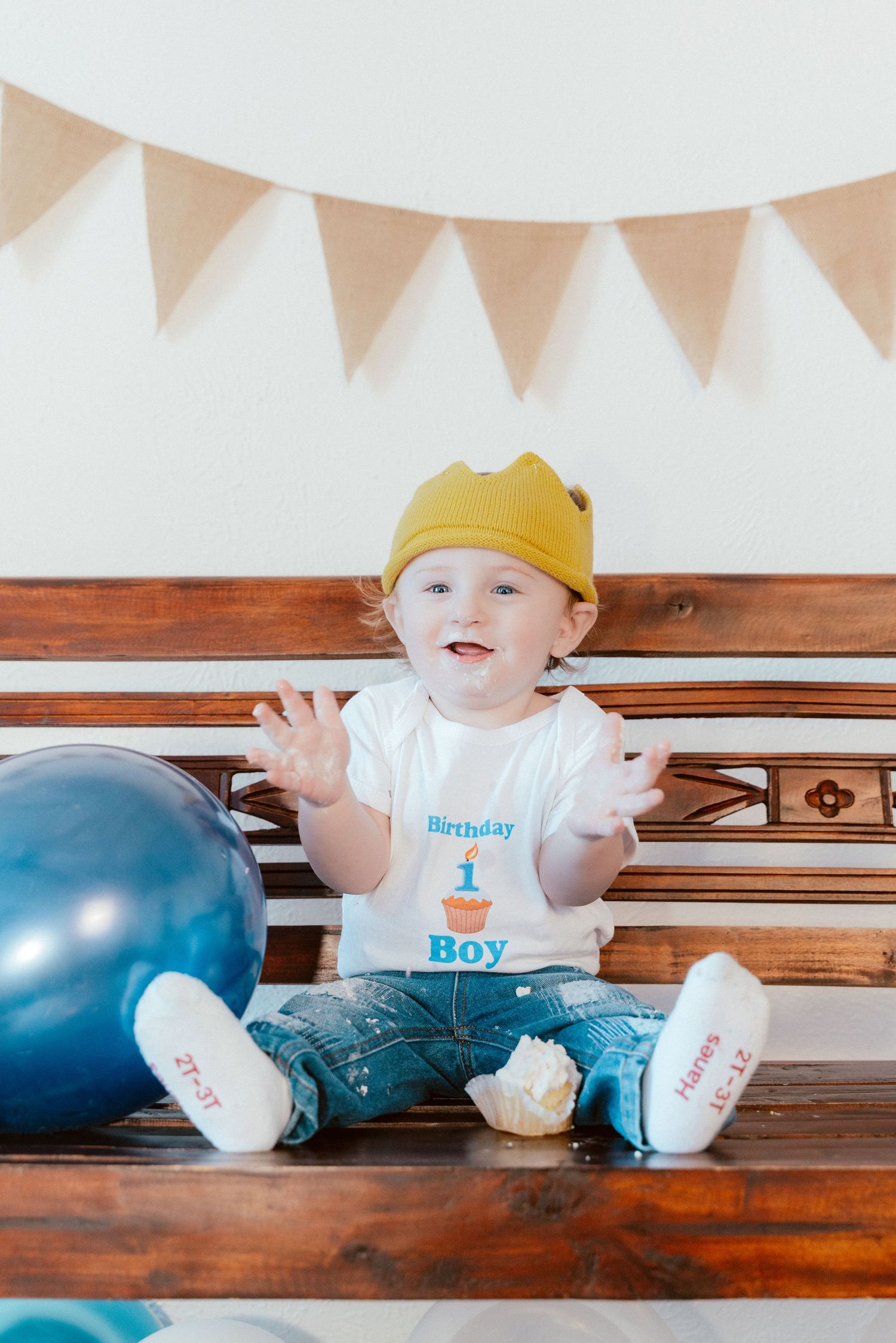 First birthday celebration with yellow crown, birthday outfit, and blue balloon on wooden bench under white bunting decorations.