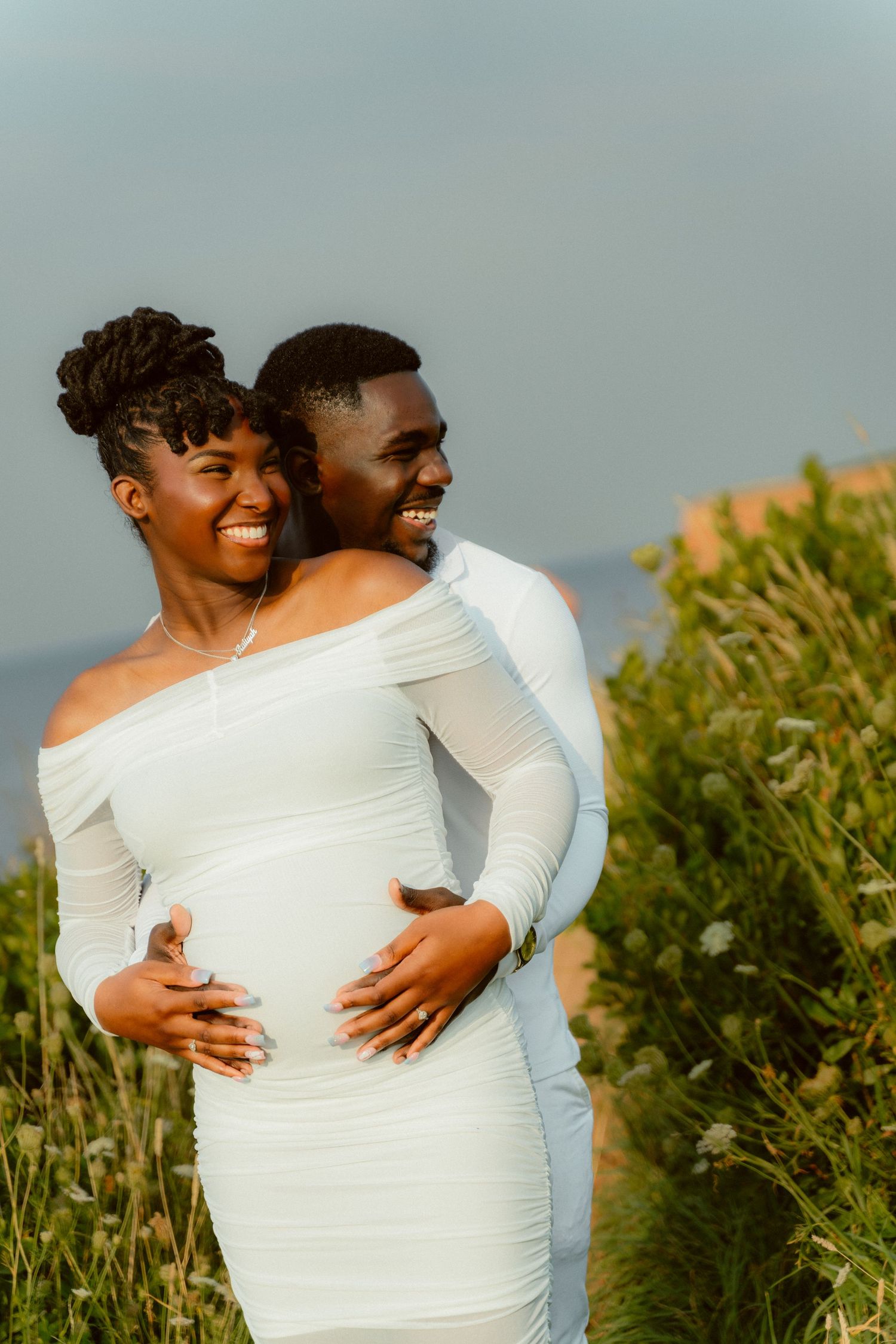 Couple in white embraces during sunset maternity photoshoot against natural backdrop.