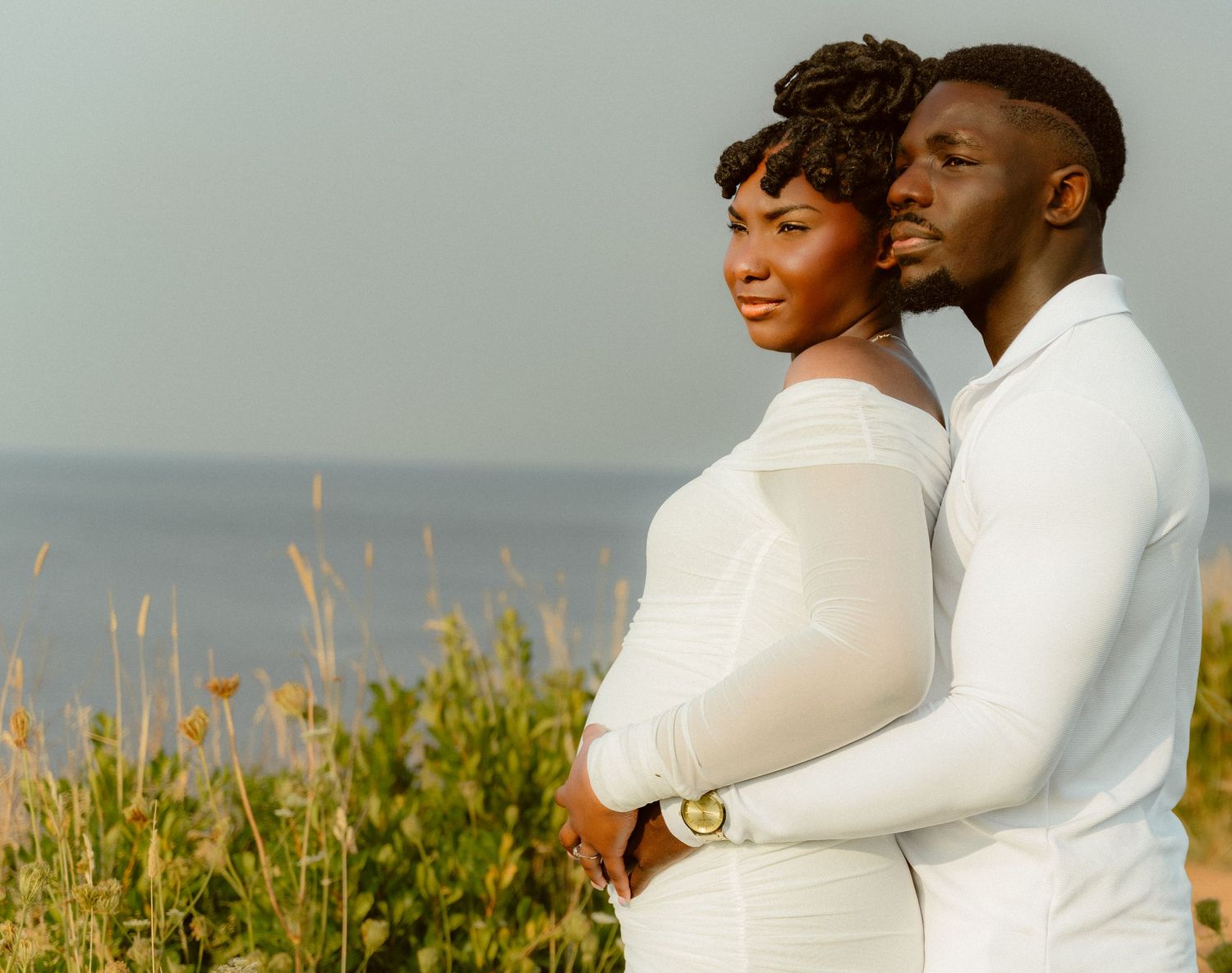 Elegant maternity portrait of couple in matching white outfits against coastal scenery.