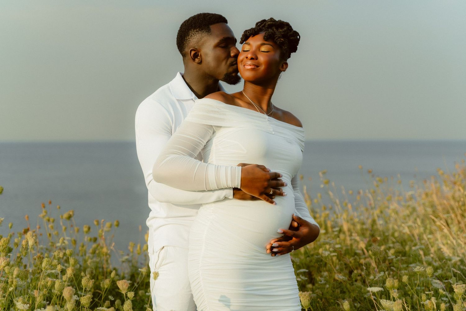 A loving couple in matching white outfits embrace near blooming wildflowers by the ocean.