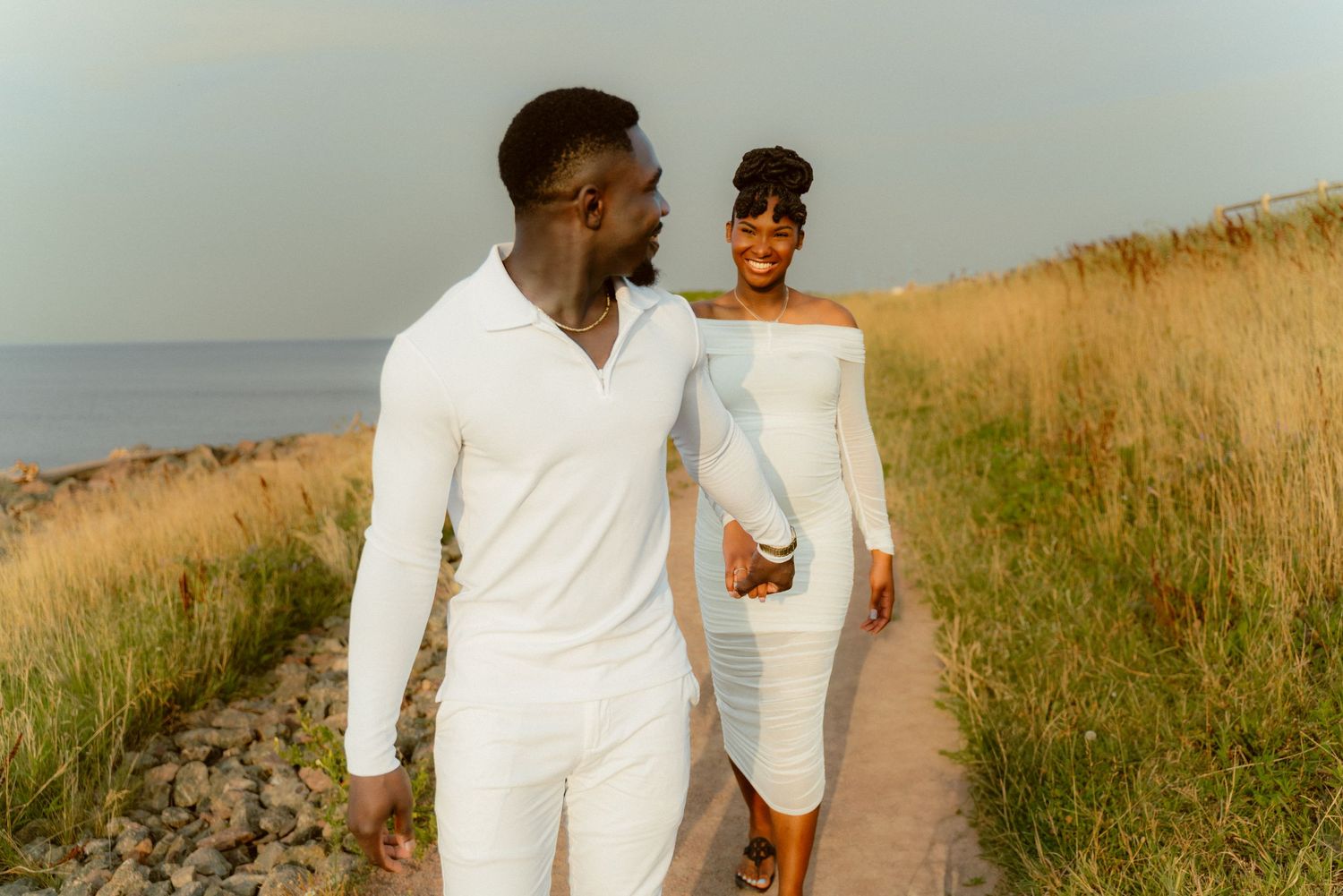 Couple in white outfits walking along a coastal path at sunset.