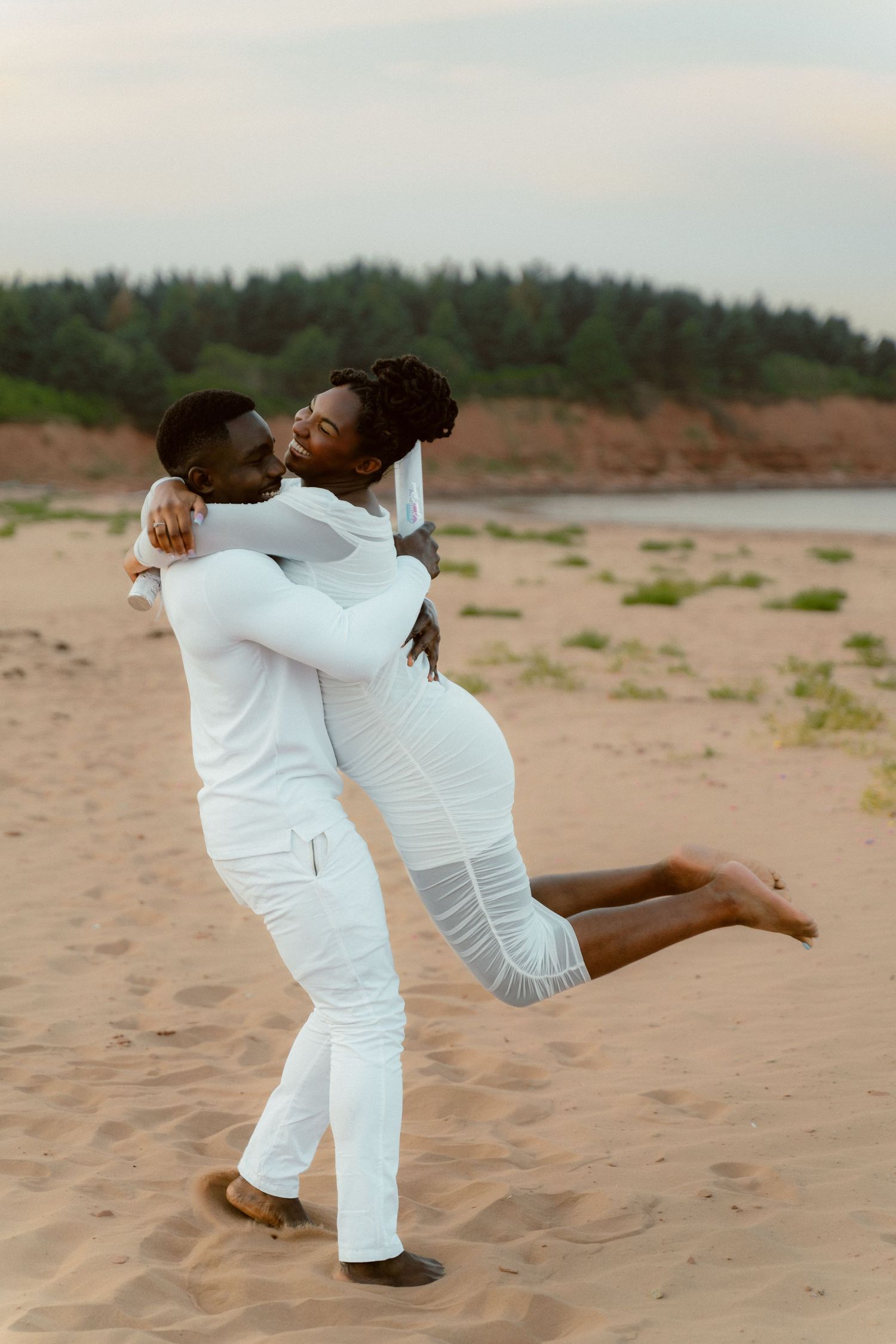 A romantic couple in white outfits sharing a joyful embrace on a sandy beach.