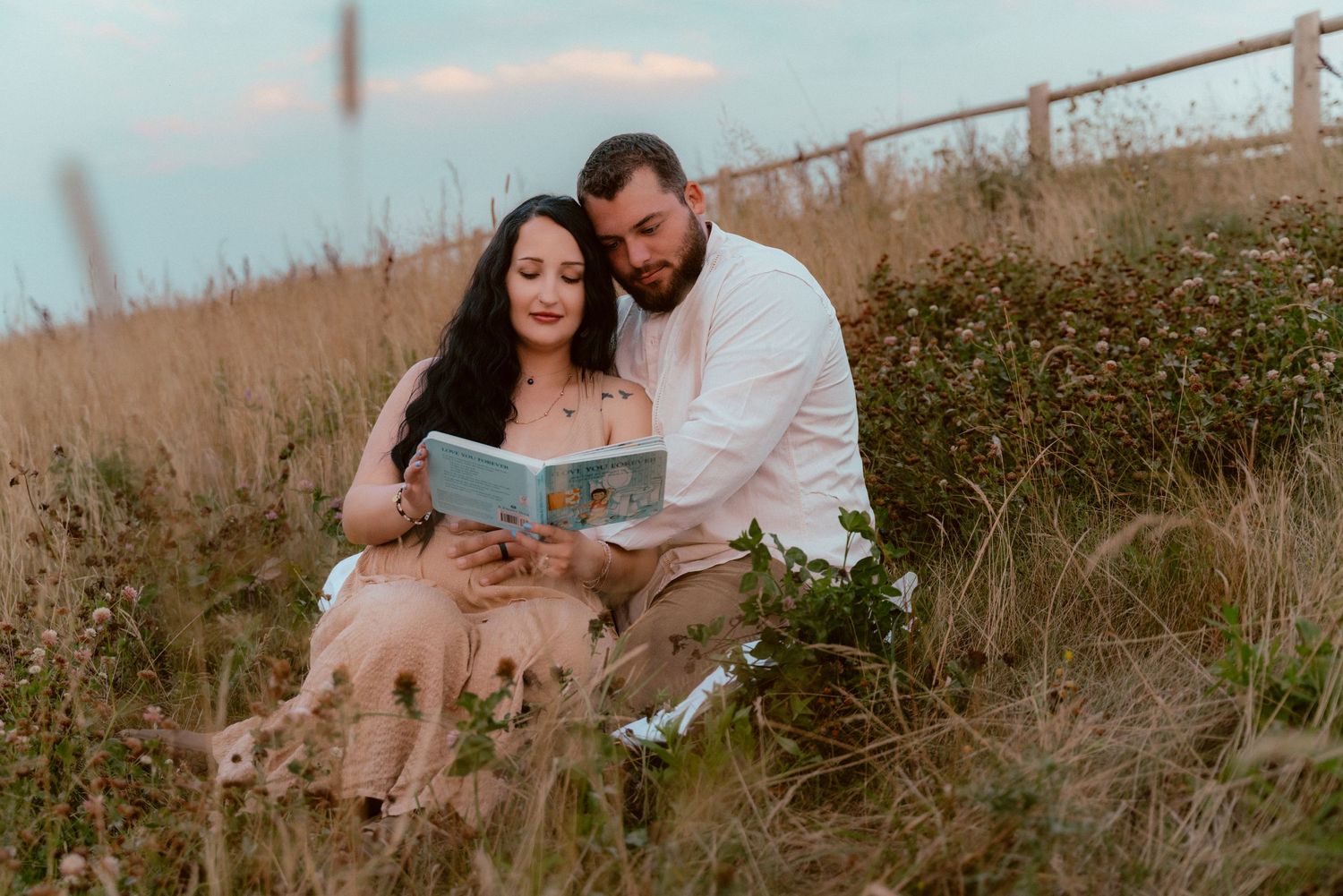 A couple sits in tall grass looking at a book together during sunset.