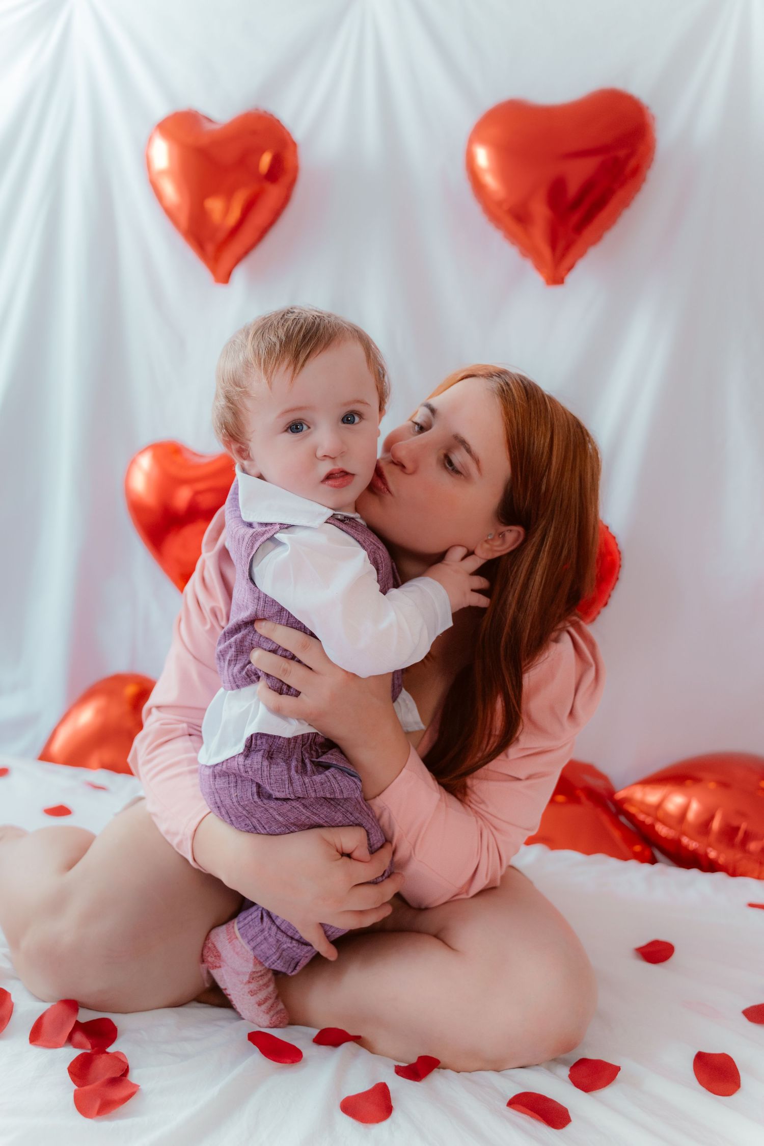 A sweet Valentine's Day portrait against white backdrop with red heart balloons and scattered rose petals.
