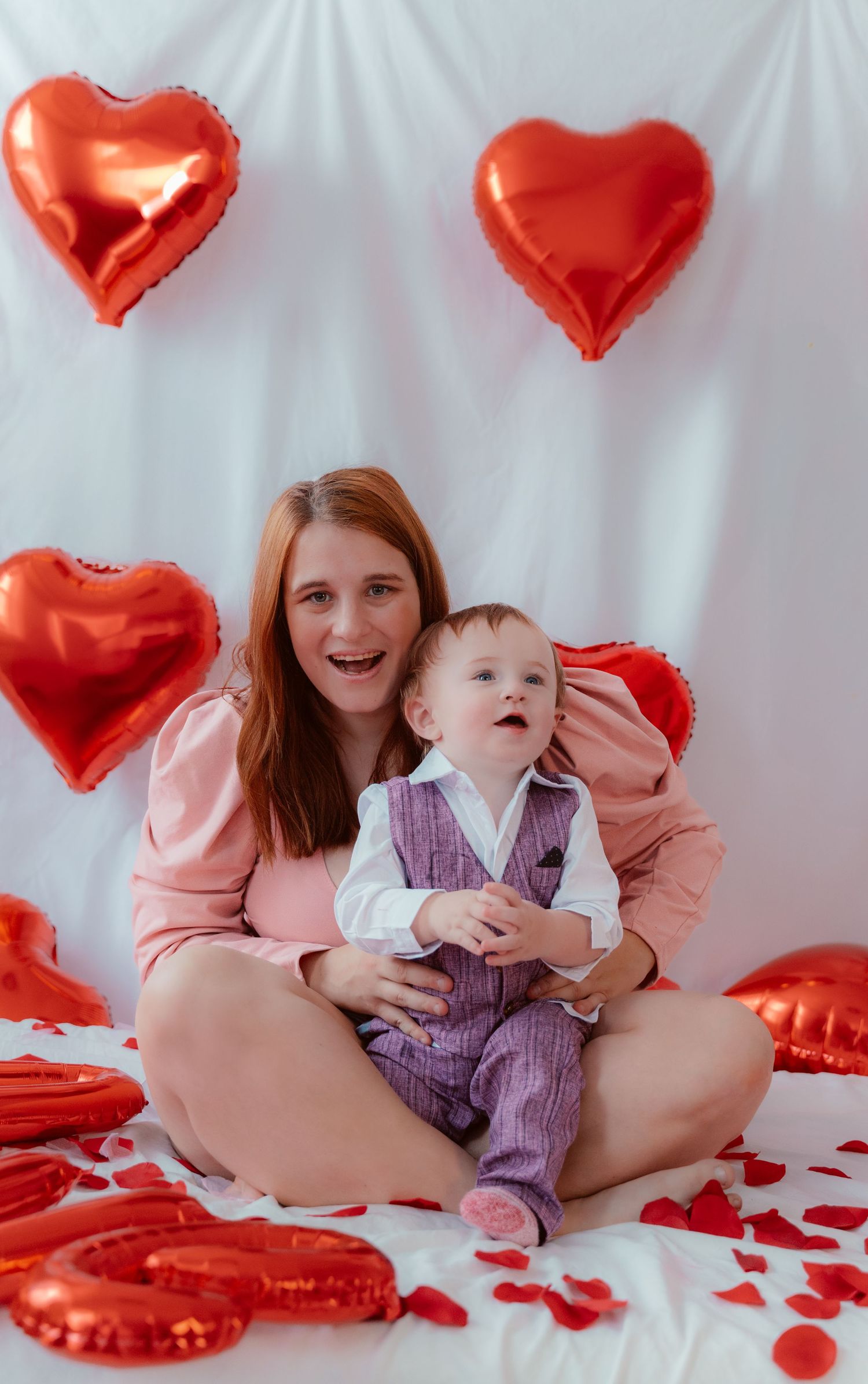 Joyful holiday portrait surrounded by red heart balloons and rose petals on white background.