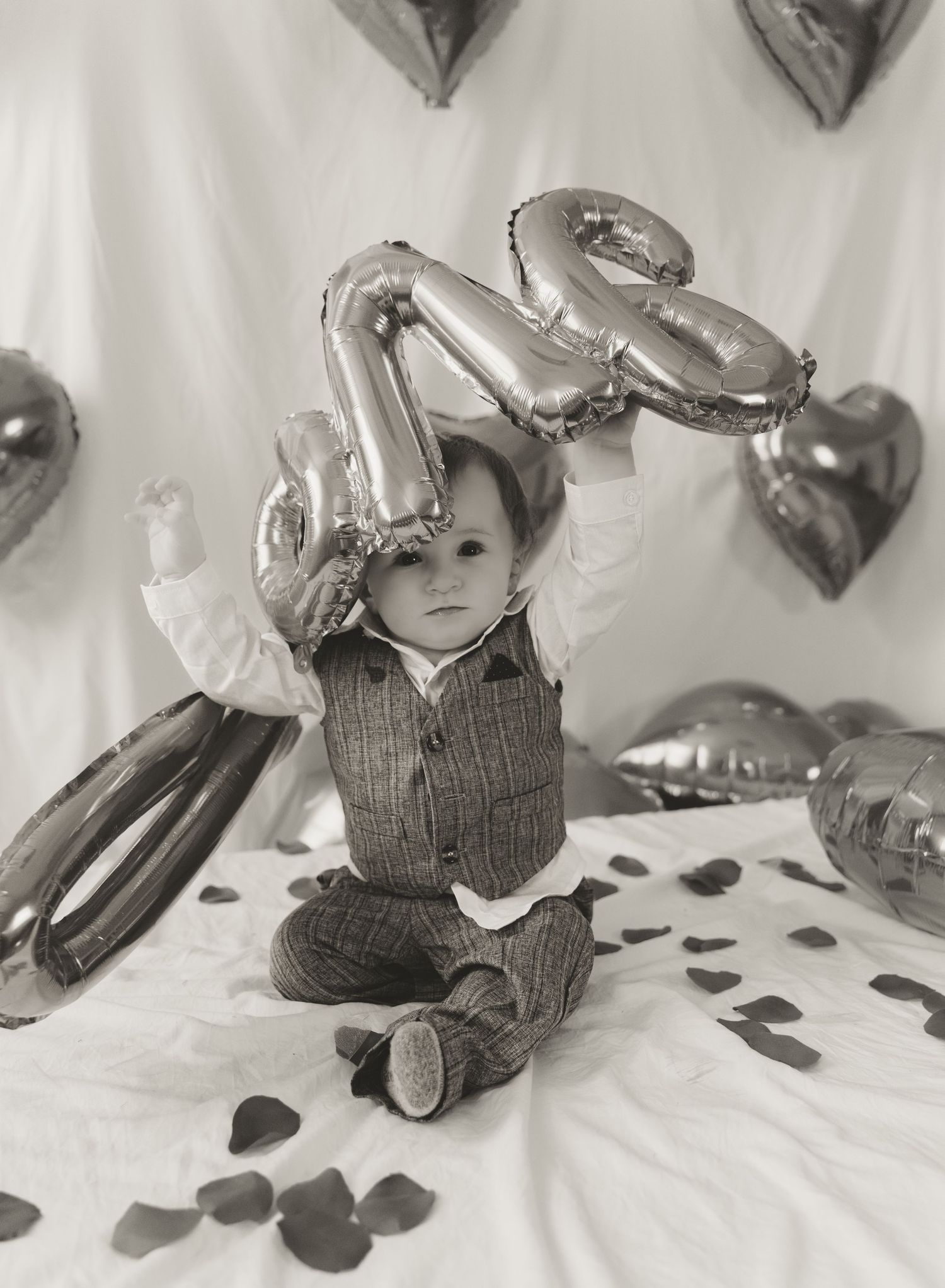 Black and white photo of a toddler sitting with silver number balloons and heart decorations on a white background.