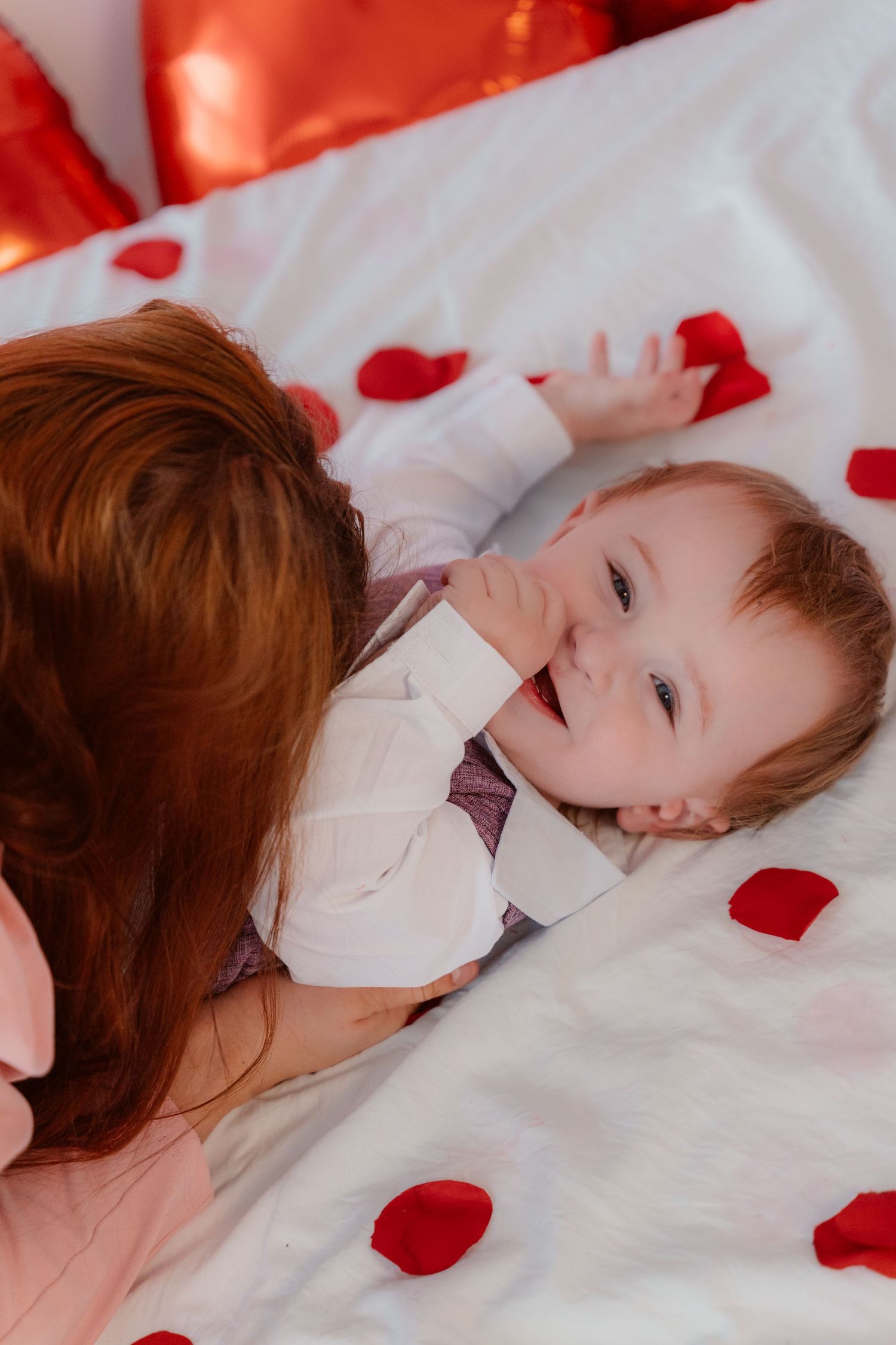 Small child laying on white bedding decorated with red rose petals while being comforted.