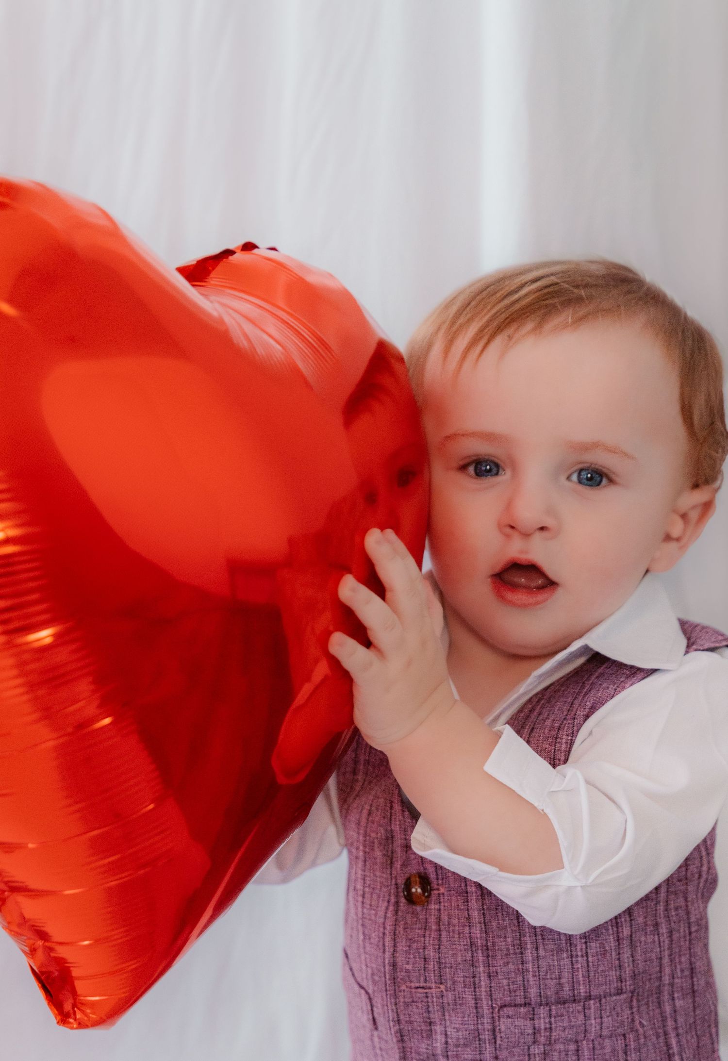 A young child holds a large red heart-shaped balloon against a light-colored background.