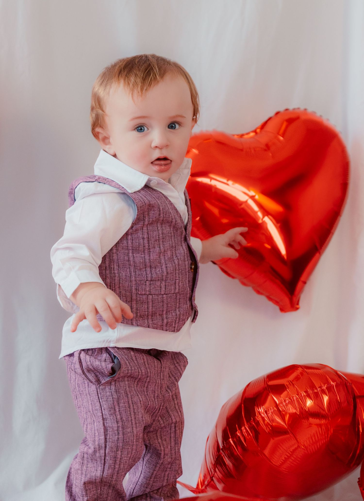 A small child in a purple vest and pants outfit stands next to two red heart-shaped Valentine's Day balloons.