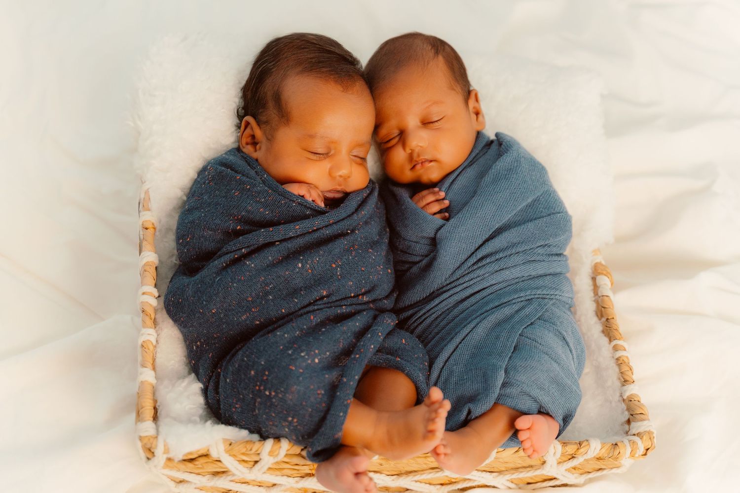 Two sleeping newborns swaddled in denim blue blankets rest peacefully together in a woven basket.