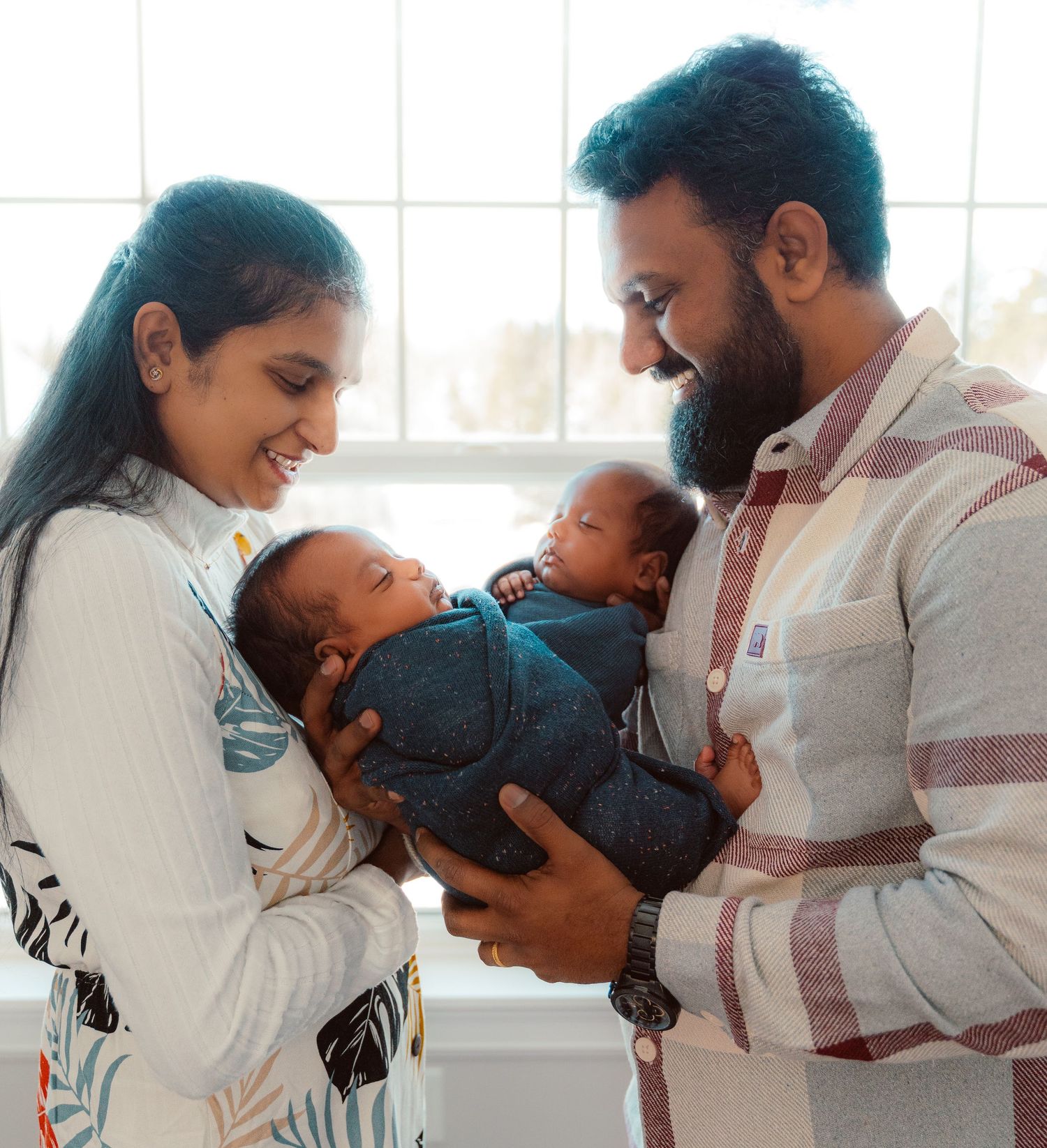 Parents holding and lovingly gazing at their twin babies near a bright window in a tender family moment.
