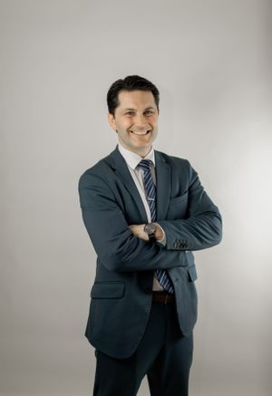 Professional headshot of a person in a navy suit with arms crossed against gray background.