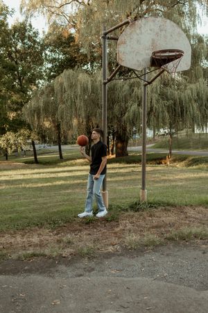 Young man in black shirt holds basketball near rustic outdoor hoop in park.