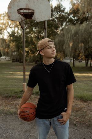 Young man in black tee and tan cap holds Spalding basketball on outdoor court with hoop and trees in background.