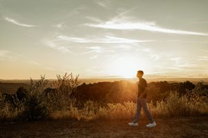 Person walking at golden hour sunset in a vast open field with warm glowing light and wispy clouds in the sky.
