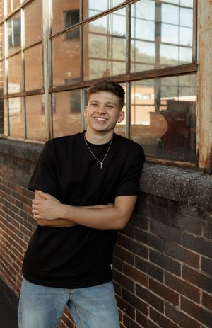 Young man in black t-shirt and jeans smiling with arms crossed, leaning against a rustic brick wall with industrial windows.