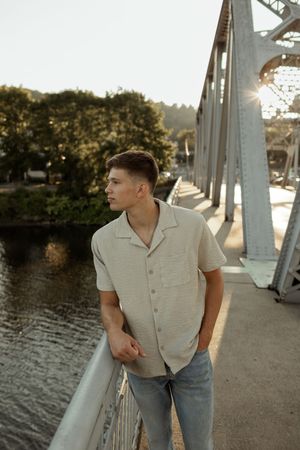 Young man in beige shirt and jeans leaning on bridge railing with river and trees in golden hour sunlight.