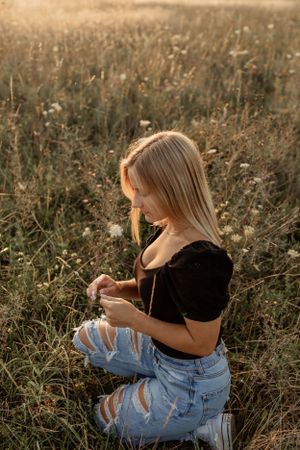 Person sitting in tall grass field wearing distressed denim and black top during golden hour.