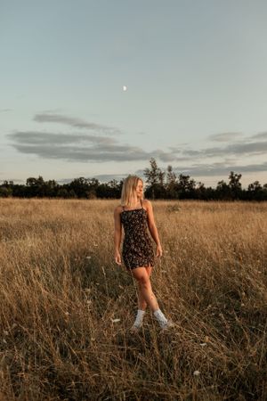 Person in floral dress standing in tall grass field at dusk with moon visible.