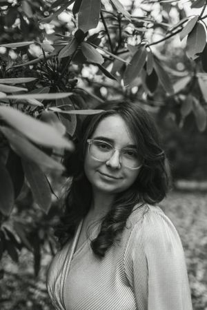 Black and white portrait of someone in glasses near foliage with natural lighting.