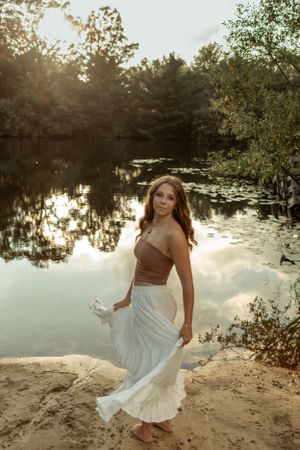 Ethereal sunset portrait by a reflective pond wearing a flowing white skirt and brown top.