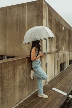 Figure with clear umbrella stands against concrete wall on rainy day wearing light denim jeans.