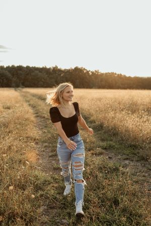 Woman walking through golden field at sunset wearing ripped jeans and black top.