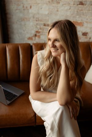 Smiling blonde woman in white outfit sitting on brown leather sofa with laptop, brick wall background.