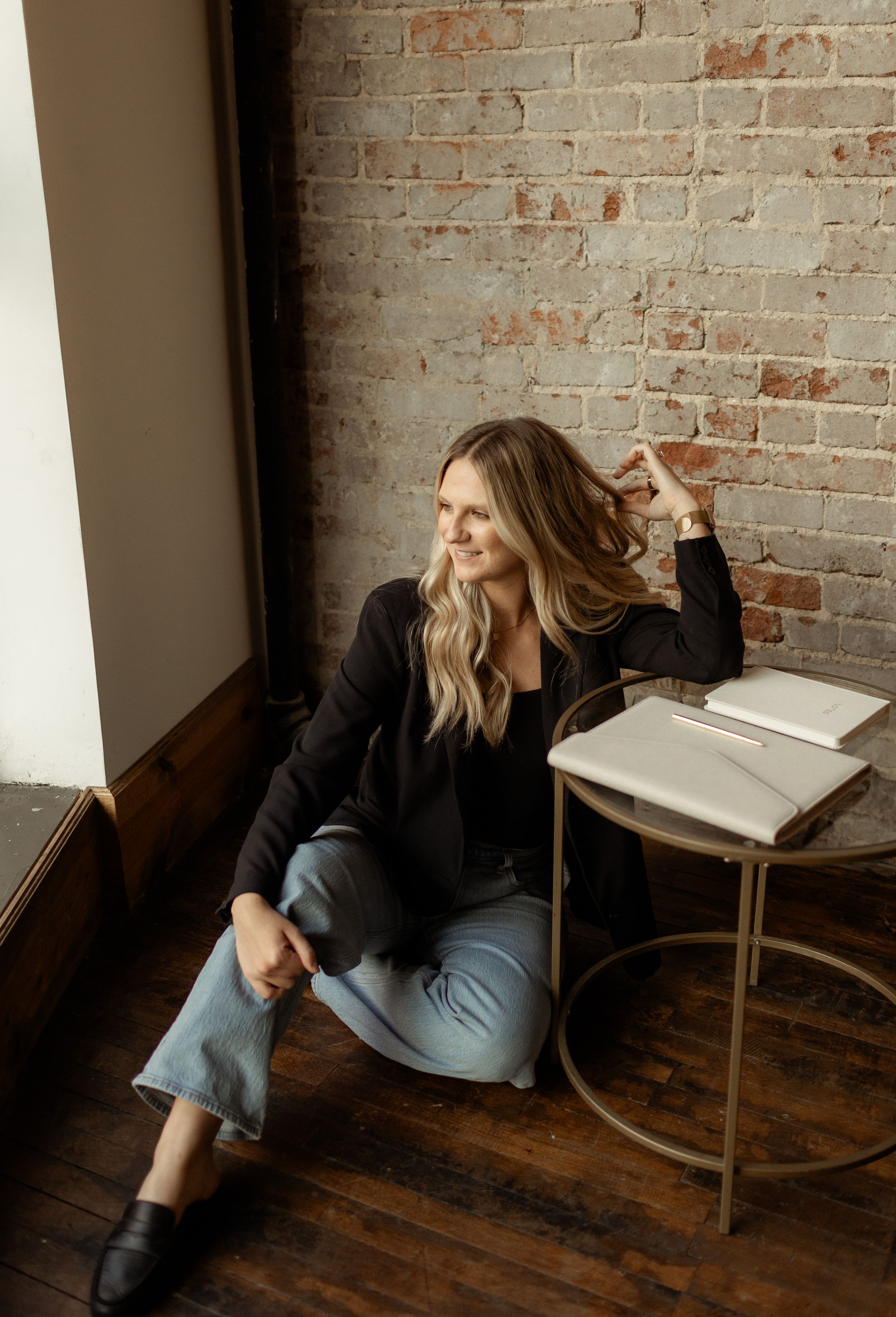 Blonde woman in black blazer sitting on wooden floor against brick wall with open book on gold side table.