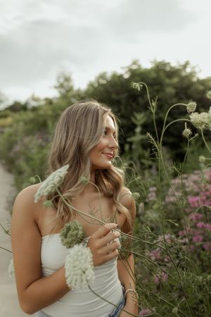 Person in white dress poses joyfully in a meadow with blooming wildflowers and natural greenery at golden hour.