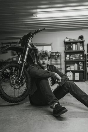Young man sitting on garage floor leaning against a dirt bike, black and white moody portrait photography.