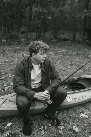 Young man sitting on a Field & Stream kayak outdoors, holding a fishing rod, in a black and white photo.