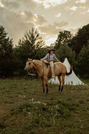 A rider on a palomino horse stands in a grassy field at sunset with a white tent in the background and dramatic clouds above.