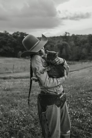 A cowboy in Western attire shares a tender moment with a black cat in a rural field under cloudy skies.