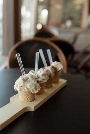 Four ice cream cones in waffle cups on a wooden board, each topped with creamy scoops and spoons, displayed on a dark table.