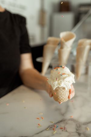 Hand holding a waffle cone with white ice cream and colorful sprinkles at a marble counter ice cream shop.