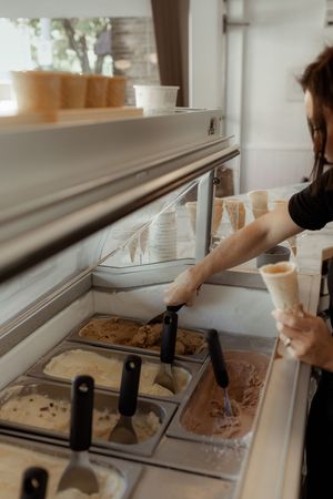 Ice cream shop worker scooping gelato flavors into a waffle cone at a commercial display freezer.