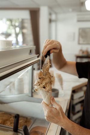 Person scooping creamy ice cream into a waffle cone at an ice cream shop counter.