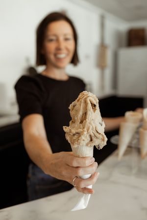 Smiling woman in black shirt holding out a waffle cone with a generous scoop of coffee ice cream in a shop.