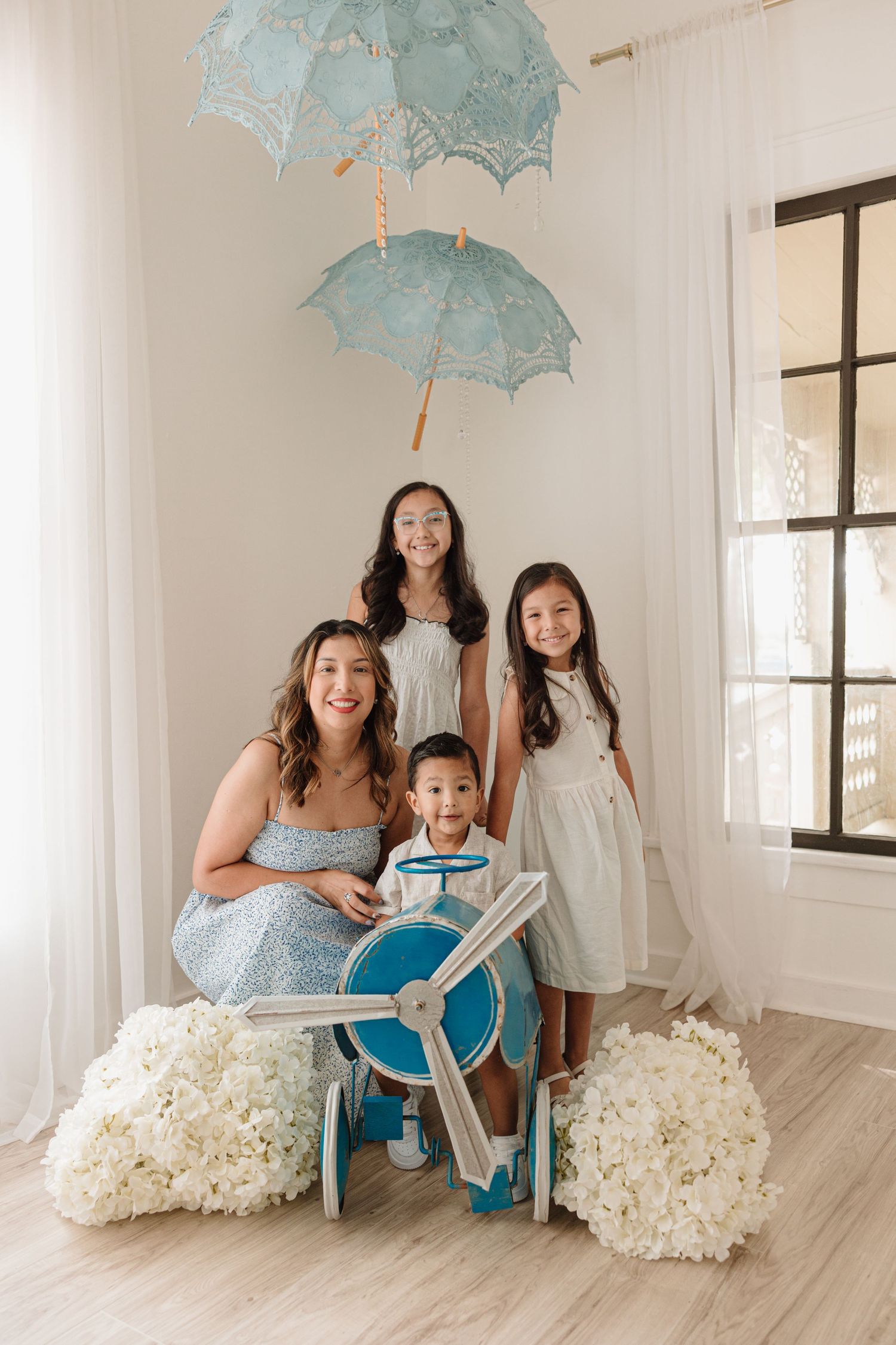 A bright indoor portrait setup with pale blue parasols, white floral decorations, and a blue toy airplane prop.