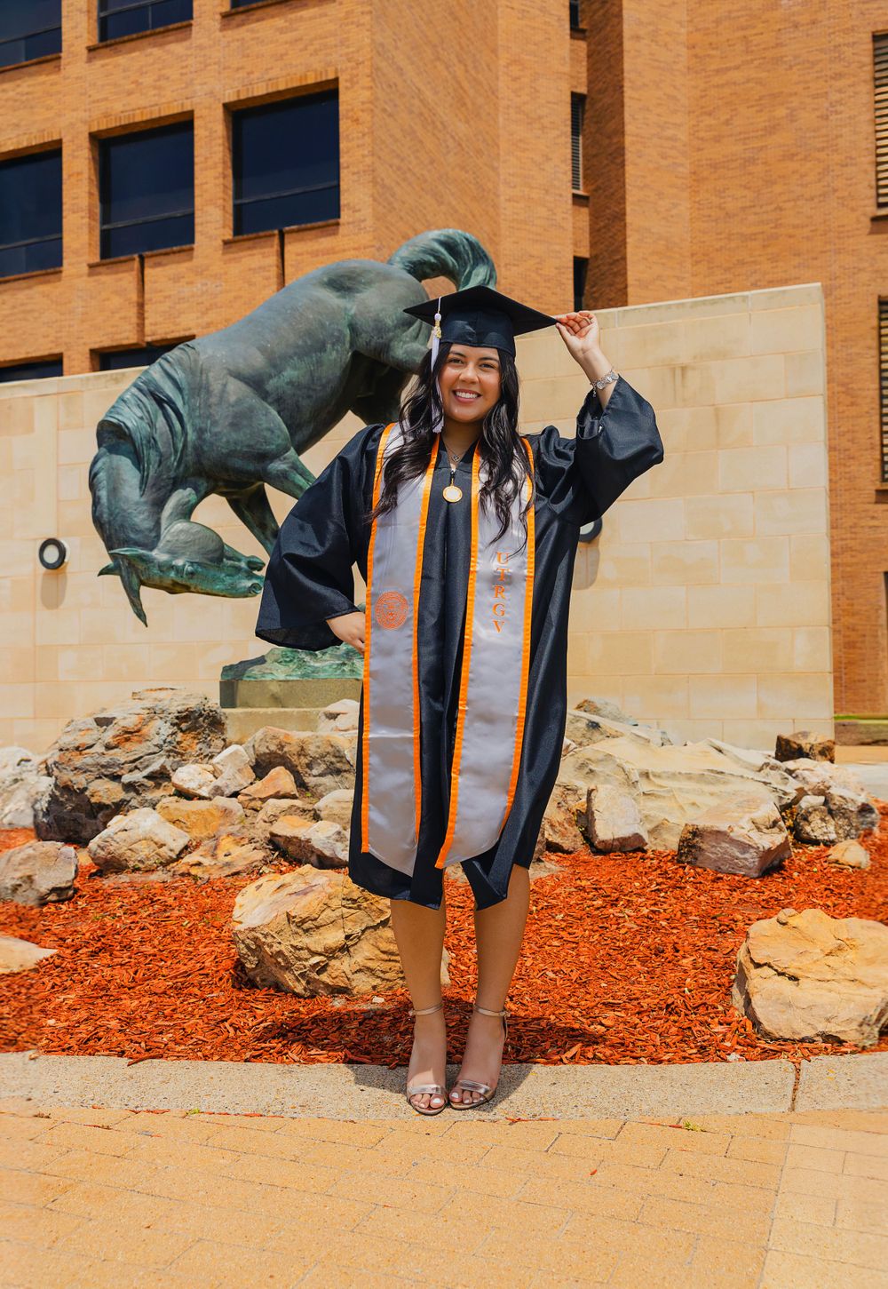 Graduate in cap and gown poses playfully with dinosaur statue on campus grounds.